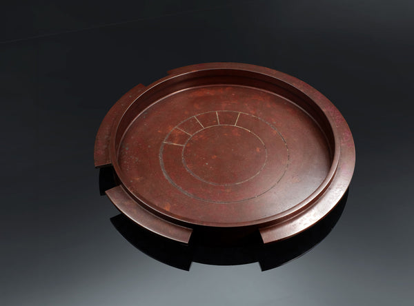 A round reddish-brown cast bronze tray with a segmented rim and engraved concentric circle details, displayed on a dark reflective surface.