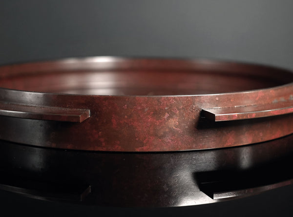 A close-up of a round reddish-brown cast bronze tray with a segmented rim and engraved concentric circle details, displayed on a dark reflective surface.
