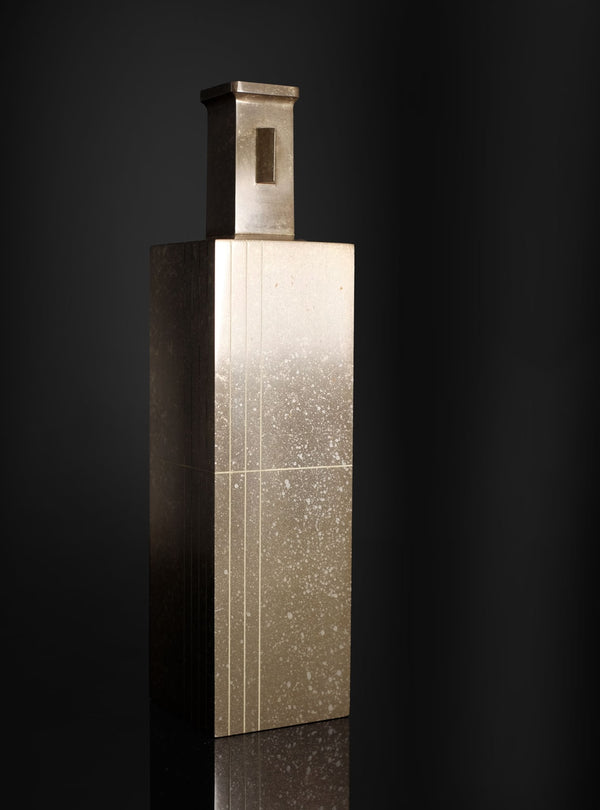 A modernist rectangular metal sculpture with a textured, speckled finish, featuring a vertical slot on its top cap, set against a dark reflective background.
