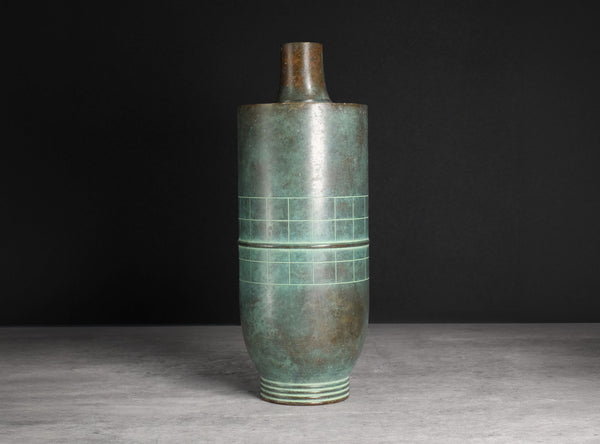 An antique-style cylindrical bronze vase with a green patina and geometric grid engravings, set against a dark gray background.