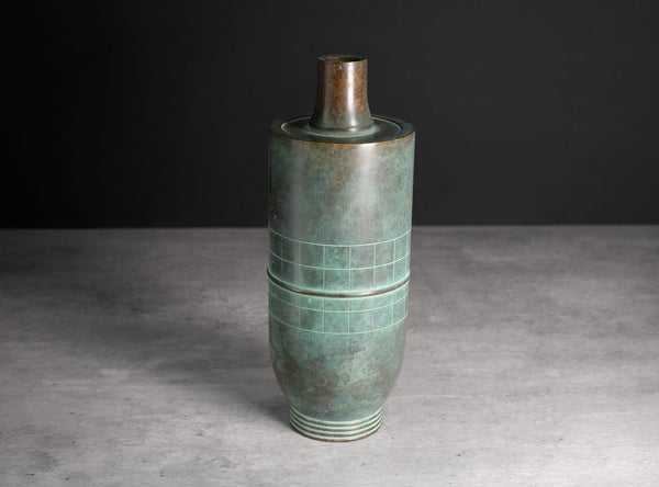 An antique-style cylindrical bronze vase with a green patina and geometric grid engravings, set against a dark gray background.