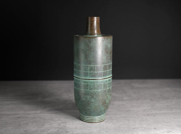 An antique-style cylindrical bronze vase with a green patina and geometric grid engravings, set against a dark gray background.