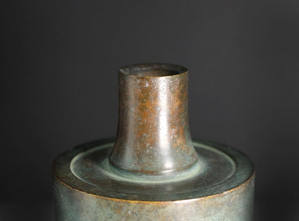 A close-up of an antique-style cylindrical bronze vase with a green patina and geometric grid engravings, set against a dark gray background.