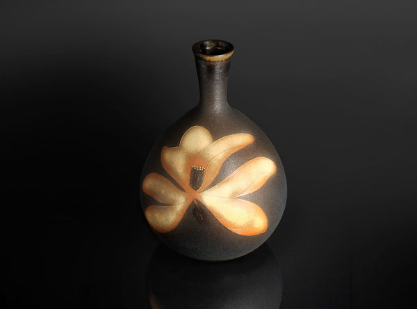 A dark brown ceramic vase with a rounded body and a short neck, decorated with a large beige floral motif on the front, set against a dark reflective background.