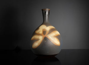 A dark gray ceramic vase with a matte finish, featuring a large, stylized flower motif in warm beige and light brown tones on its front, set against a dark gray background.