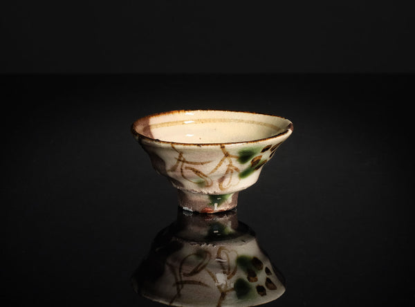 A small ceramic footed bowl with a cream-colored glaze, decorated with brown floral patterns and green accents, set against a dark gray gradient background with a reflective surface below.