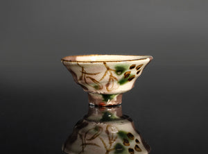 A small ceramic footed bowl with a cream-colored glaze, decorated with brown floral patterns and green accents, set against a dark gray gradient background with a reflective surface below.