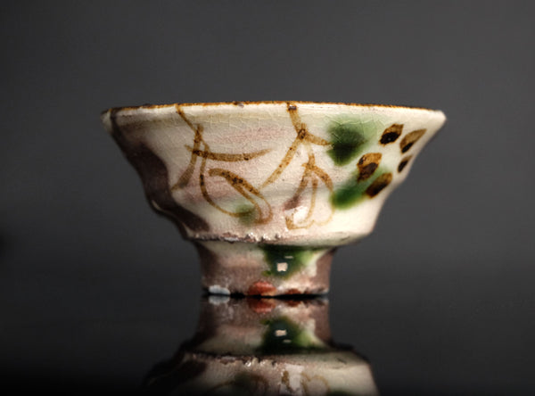 A handmade ceramic footed bowl with a textured off-white glaze, decorated with brown brushstrokes and green accents, set against a dark gray background with a soft reflection below.