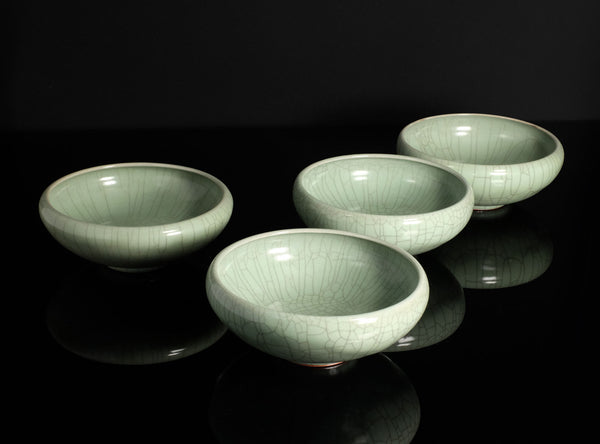 Four light green celadon ceramic bowls with a crackle glaze, arranged on a reflective black surface against a dark background.
