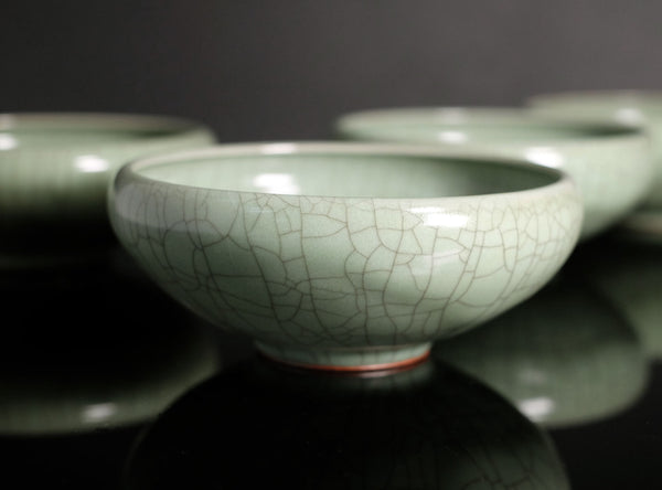 A close-up of a glossy pale green ceramic bowl with a crackle glaze pattern, shown with other matching bowls blurred in the background on a dark reflective surface.