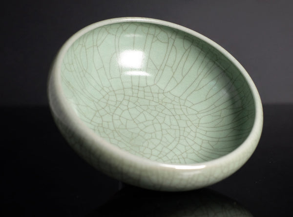 A ceramic bowl with a pale celadon green glaze and a prominent crackle pattern, shown on a dark reflective surface.