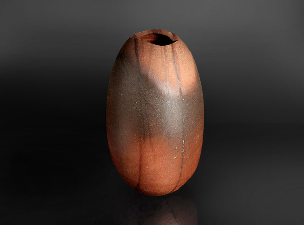 A small organic-shaped ceramic vase with a wide opening, featuring a mottled finish that transitions from dark brown to warm terracotta orange, set against a dark gradient background.