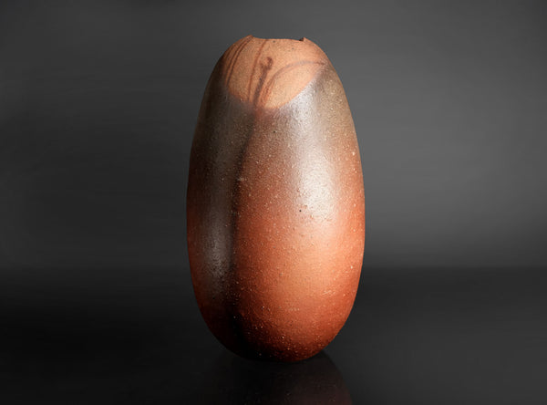 A small organic-shaped ceramic vase with a wide opening, featuring a mottled finish that transitions from dark brown to warm terracotta orange, set against a dark gradient background.
