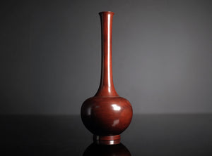 A glossy reddish-brown cast bronze vase with a bulbous base and a long, slender neck, standing on a dark reflective surface against a dark gray background.