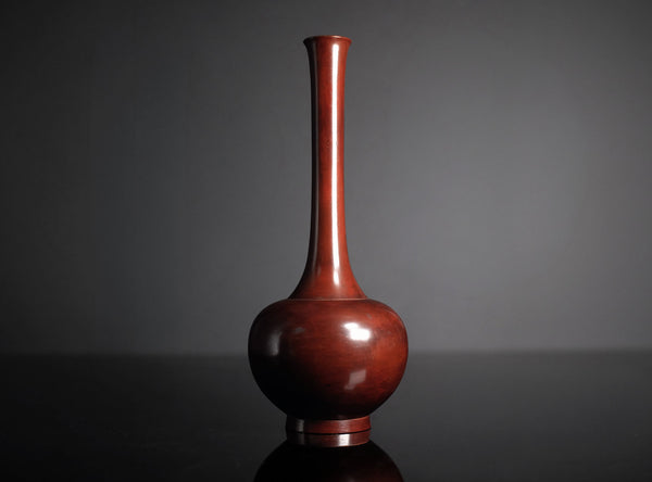 A glossy reddish-brown cast bronze vase with a bulbous base and a long, slender neck, standing on a dark reflective surface against a dark gray background.