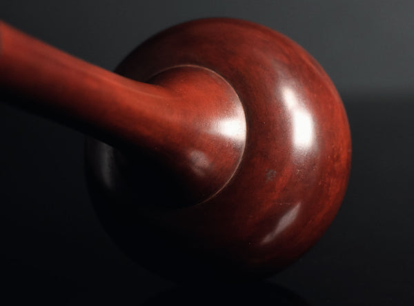 A glossy reddish-brown cast bronze vase with a bulbous base and a long, slender neck, standing on a dark reflective surface against a dark gray background.
