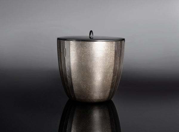 A metallic cylindrical container with a ribbed textured surface and a matching lid featuring a small loop handle, set against a dark gray gradient background with a reflective black surface below.