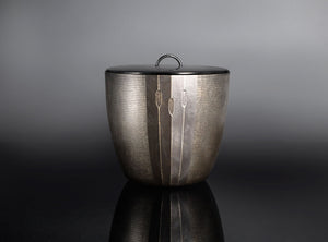 A textured silver metal container with a matching lid and a small top loop handle, photographed on a dark reflective surface against a gray gradient background.