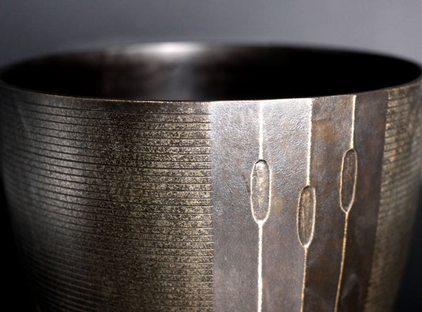 Close-up of a dark metallic bowl with horizontal grooved texture and three vertical oval cutouts on the front, shown against a dark gray background.