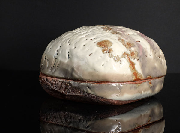 A handmade ceramic lidded box with a textured, organic-shaped cream and brown glazed top and a rustic dark brown base, displayed on a reflective black surface against a black background.
