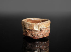 A rustic, handcrafted ceramic cup with a rough, textured surface resembling natural stone, featuring earthy brown, beige, and reddish tones, set against a dark reflective background