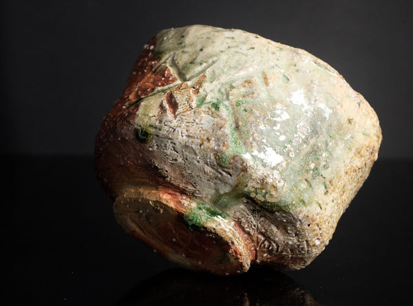 A handmade ceramic bowl with a rough, organic shape and a textured glaze featuring green, beige, and brown tones, photographed on a reflective black surface.