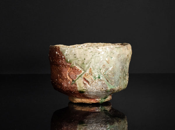 A handmade ceramic bowl with a rustic, hand-painted glaze featuring a mix of brown, green, and off-white colors, displayed on a reflective black surface against a dark background.