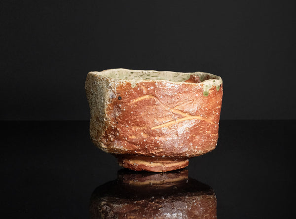 A handmade ceramic tea bowl with a textured, rustic finish, featuring a reddish-brown glazed exterior and a light beige interior, set against a solid black background with a reflective surface below.