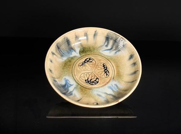 A ceramic plate with a beige and green glaze, featuring blue painted brushstrokes around the rim and a central black and green floral motif, displayed on a black background.