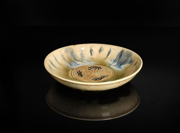 A ceramic shallow plate with a beige and blue glaze, featuring hand-painted black calligraphy in the center, displayed on a reflective black surface against a black background.