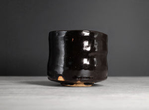 A handmade dark brown glazed ceramic cup with a glossy finish and a small exposed clay foot, set against a dark gray background on a light gray surface.