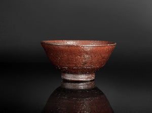 A handmade ceramic tea bowl with a textured, earthy reddish-brown glaze, shown on a reflective dark surface against a solid black background.