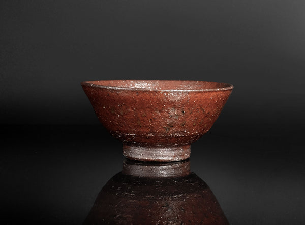 A handmade ceramic tea bowl with a textured, earthy reddish-brown glaze, shown on a reflective dark surface against a solid black background.