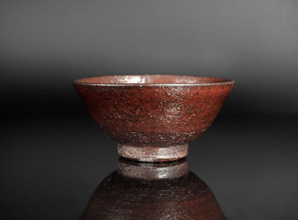 A handmade ceramic tea bowl with a textured, earthy reddish-brown glaze, shown on a reflective dark surface against a dark gray background.