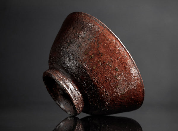 A dark brown textured ceramic tea bowl with a glossy glaze, shown on a reflective black surface against a dark gray background.
