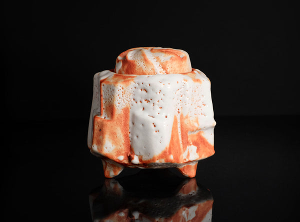 A sculptural ceramic vessel with a textured, ribbed surface, featuring a white glaze with orange and reddish-brown marbled accents, set against a solid black background with a reflective surface below.