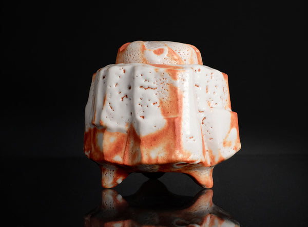 A sculptural ceramic vessel with a textured, ribbed surface, featuring a white glaze with orange and reddish-brown marbled accents, set against a solid black background with a reflective surface below.