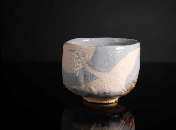 A handmade ceramic tea bowl with a textured, speckled glaze in shades of light blue-gray and warm cream, set against a solid black background with a soft reflection below.
