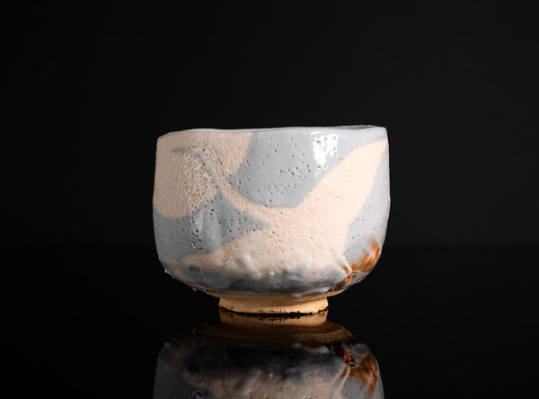 A handmade ceramic tea bowl with a textured, speckled glaze in light blue and cream, featuring a small brown foot ring, set against a solid black background with a soft reflection below.