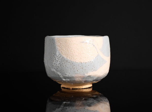 A handmade ceramic tea bowl with a textured, speckled glaze in off-white and light blue, standing on a reflective black surface against a solid black background.