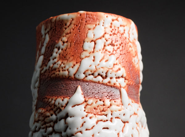 A close-up of a cylindrical ceramic vase or candle holder with a textured, crackled pattern in terracotta orange and white, featuring a dark reddish-brown horizontal band in the center, set against a dark gray background.