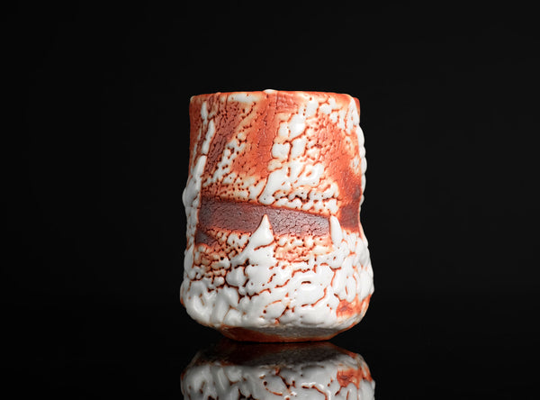 A handmade ceramic cup with a textured, organic surface pattern in white and terracotta orange, shown on a reflective black background.