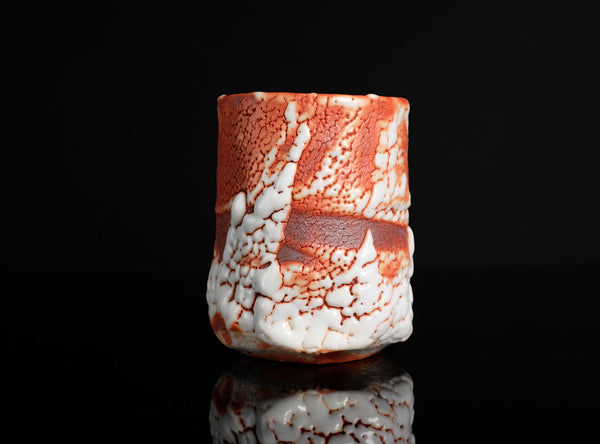 A cylindrical ceramic cup with a textured, crackled glaze pattern in terracotta orange and white, set against a solid black background with a soft reflection below.