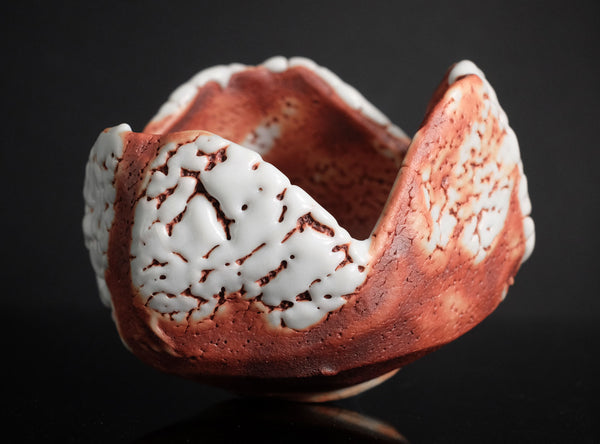 A handmade ceramic bowl with a textured, organic shape, featuring a reddish-brown clay body and white glaze splatters, set against a solid black background.