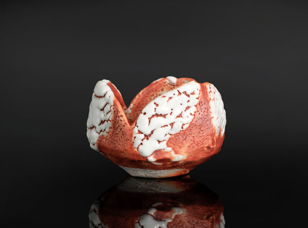 A handmade ceramic bowl with a textured, organic shape in terracotta red and white, displayed on a reflective black surface against a dark background.