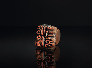 A square bronze seal stamp with red-orange engraved characters, displayed on a reflective black surface against a dark background.