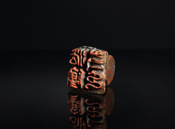 A square bronze seal stamp with red-orange engraved characters, displayed on a reflective black surface against a dark background.