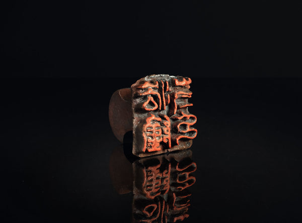 A square bronze seal stamp with red-orange engraved characters, displayed on a reflective black surface against a dark background.