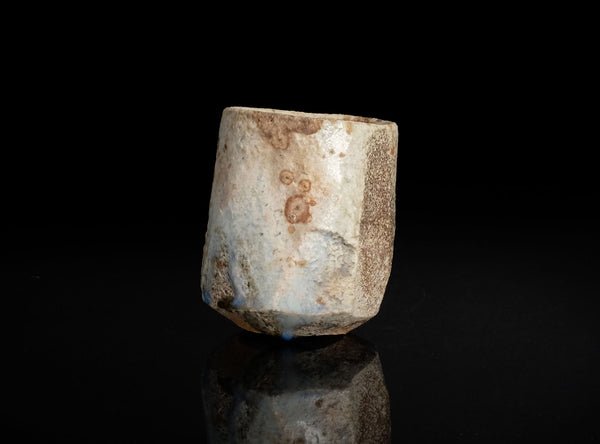 A small cylindrical stone or ceramic vessel with a rough, weathered finish in off-white and brown tones, photographed on a black reflective surface against a black background.