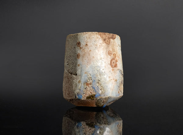 A rustic, handcrafted ceramic cup with a textured, weathered finish in earthy beige, brown, and light blue glazes, standing on a reflective black surface against a dark gray background.
